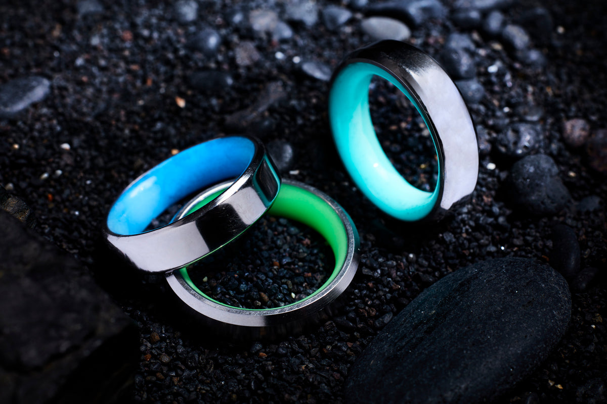 Green, Blue, and Turquoise glow rings with titanium exterior and beveled edges on a background of black rocks. 